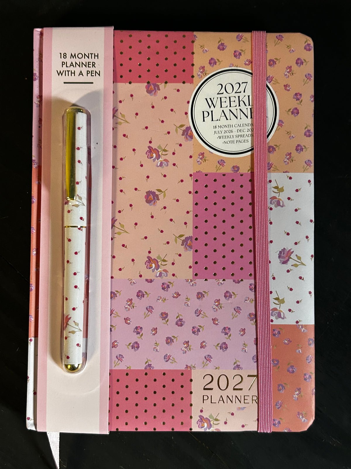A 2027 weekly planner with an elastic closure and a matching pen, featuring a patchwork floral pattern in pink, peach, and purple tones, set against a black background.