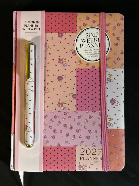 A 2027 weekly planner with an elastic closure and a matching pen, featuring a patchwork floral pattern in pink, peach, and purple tones, set against a black background.